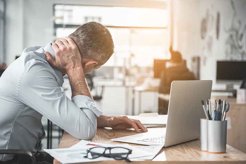 Professional Businessman holding sore neck while using notebook computer at his desk. It's said that poor posture can affect your balance - affecting your jaw along with TMJ.