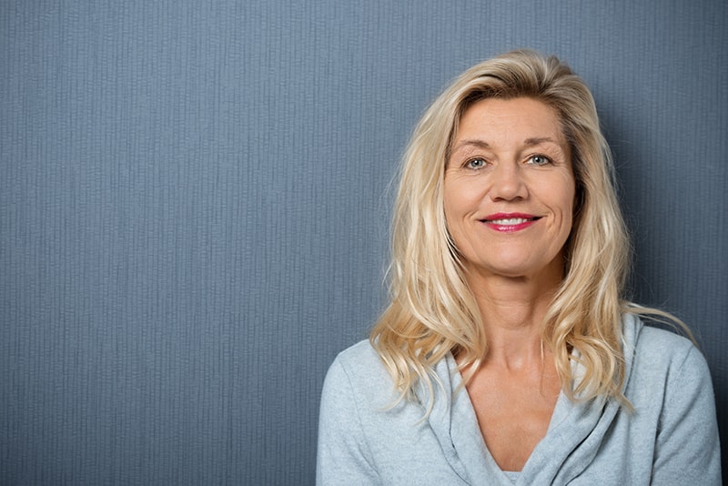 attractive older woman in front of a blue background grinning to the camera. With a smile that shines she's looking at an alternative to standard dentures with dental implants or a dental bridge.