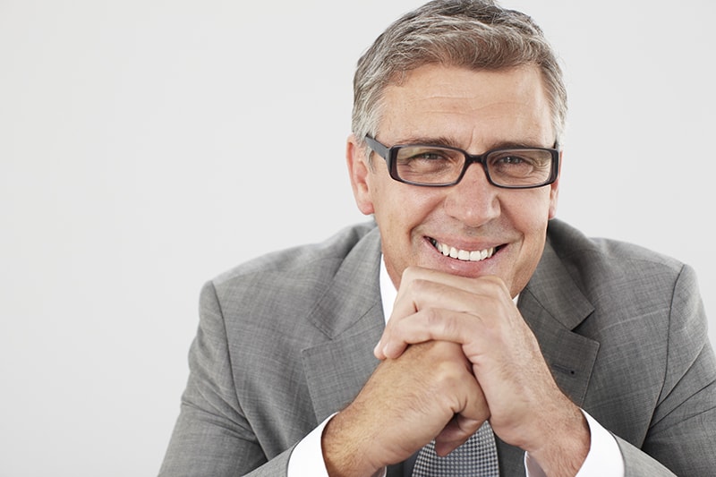 An older business man with a sincere smile, resting his hands on his chin. With the benefits of a tooth transplantation he doesn't have to have a dental implant.