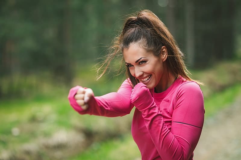 A woman in a pink workout shirt shadow boxes outside in nature. With workouts she's building her strength, with Myofunctional Therapy she's Strengthen Muscles of the Mouth and Face.