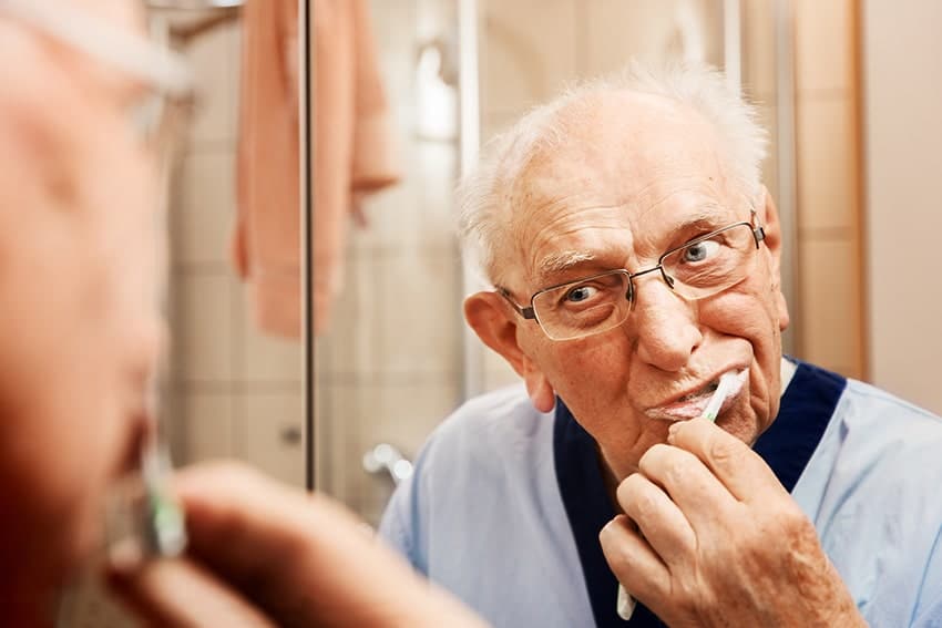 An older man brushes his teeth in front of a mirror in the bathroom